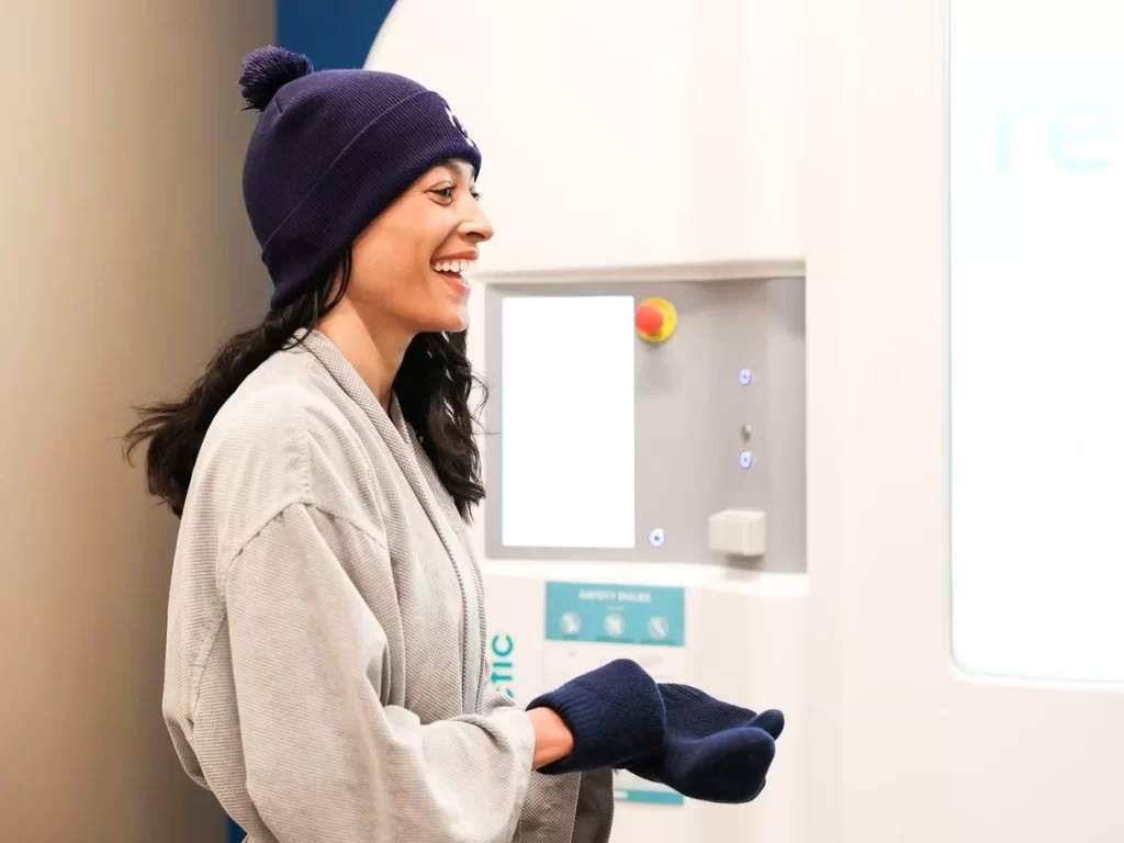 A smiling woman with a hat and gloves in front of Arctic chamber at Restore Hyper Wellness