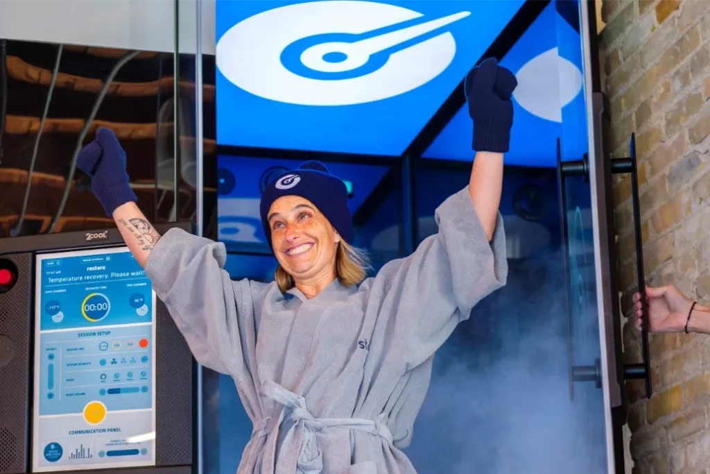 a woman with a hat a gloves smiles, as she leaves a cryochamber.