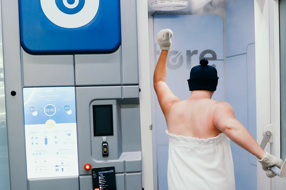 A person cheerfully entering a cryochamber while wearing gloves and a beanie hat.