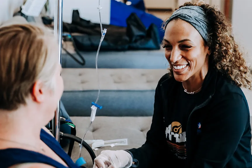 Two women smiling, one is helping the other with an IV Drip.